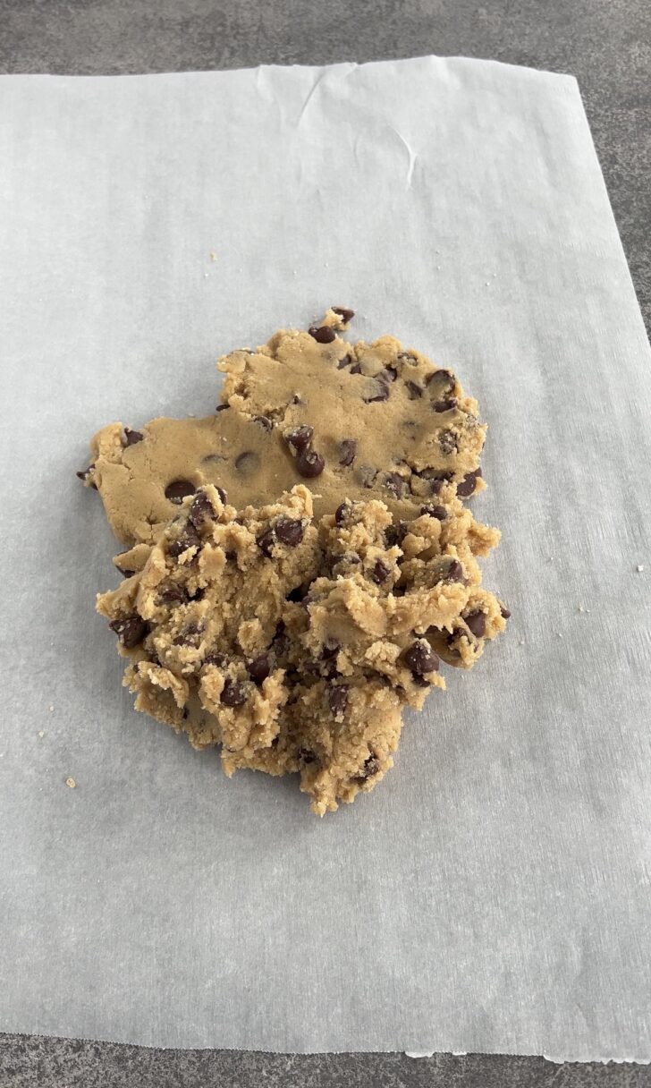 A mound of chocolate chip cookie dough sits on a sheet of parchment paper atop a gray countertop, ready to be shaped or baked.