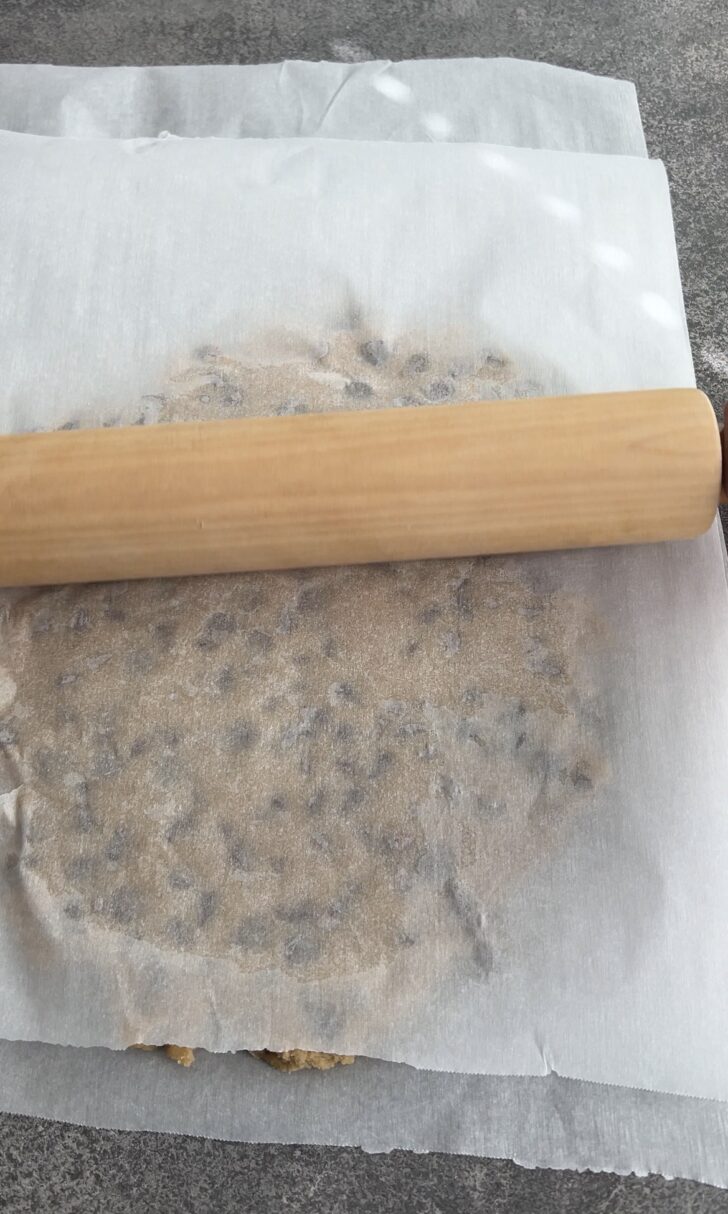 A rolling pin flattening cookie dough with chocolate chips between two sheets of parchment paper on a grey countertop.
