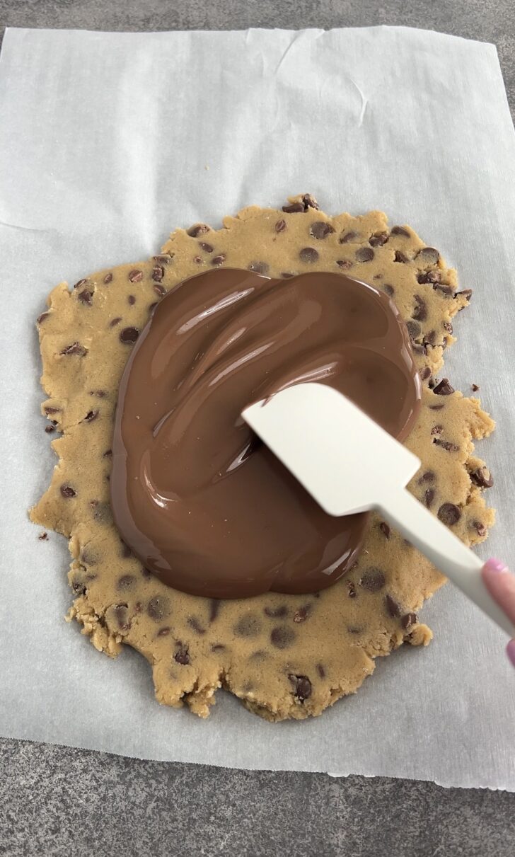 A hand spreads melted chocolate over a flattened round of chocolate chip cookie dough using a white spatula, all on parchment paper atop a gray surface.