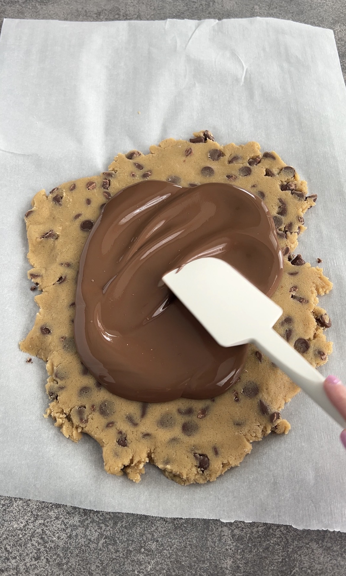 A hand spreads melted chocolate over a flattened round of chocolate chip cookie dough using a white spatula, all on parchment paper atop a gray surface.