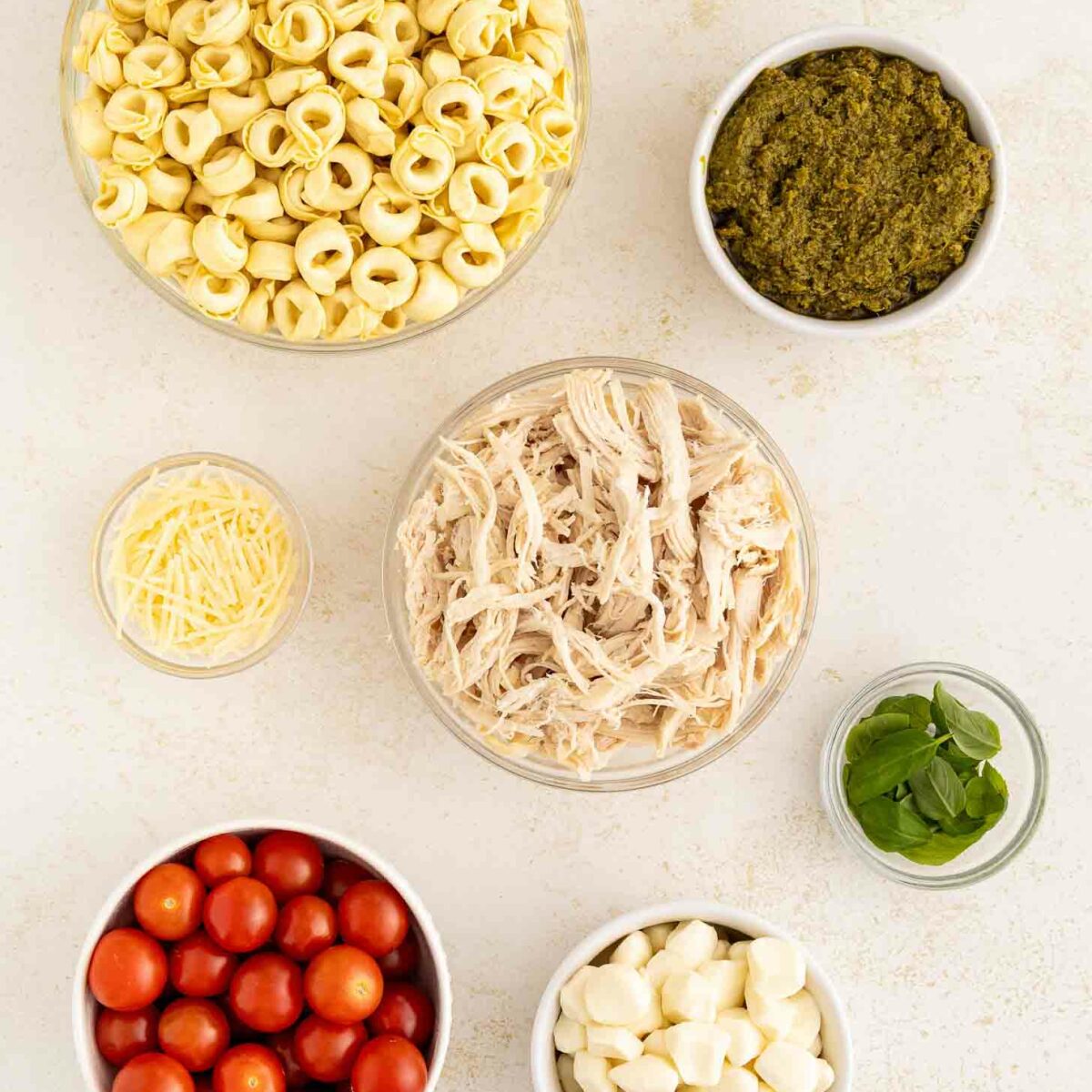 Bowls containing uncooked tortellini, shredded chicken, pesto, cherry tomatoes, mozzarella balls, grated cheese, and fresh basil are arranged on a light surface.