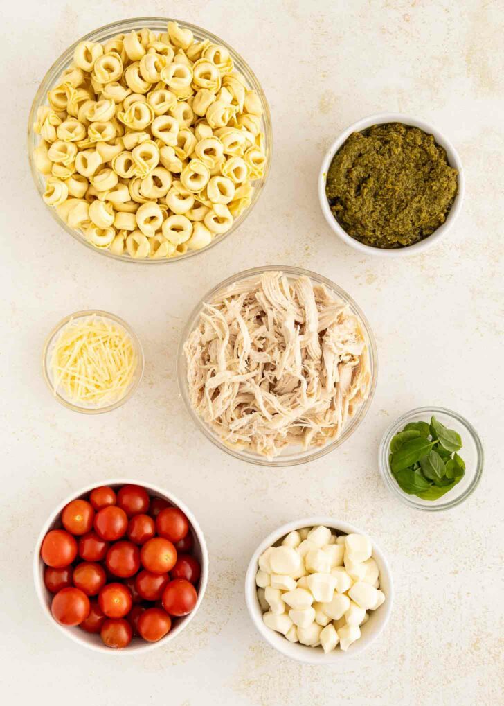 Bowls containing uncooked tortellini, shredded chicken, pesto, cherry tomatoes, mozzarella balls, grated cheese, and fresh basil are arranged on a light surface.