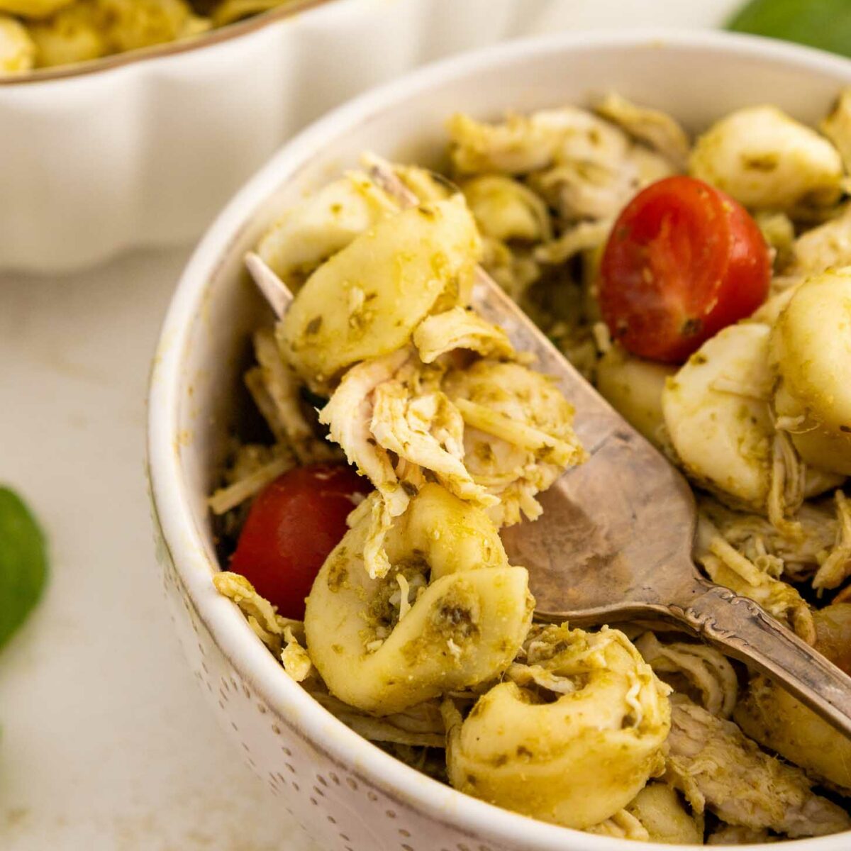 A close-up of a bowl of tortellini pasta mixed with shredded chicken, cherry tomatoes, and pesto sauce. A fork is resting inside the bowl, and another bowl of pasta is partially visible in the background.