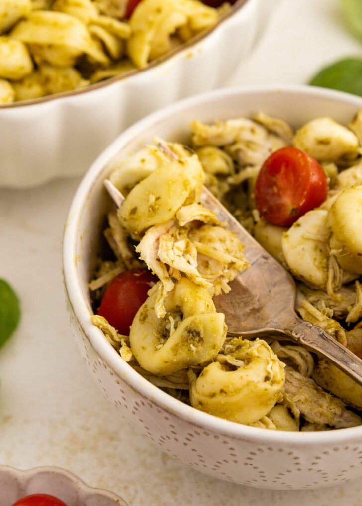 A close-up of a bowl of tortellini pasta mixed with shredded chicken, cherry tomatoes, and pesto sauce. A fork is resting inside the bowl, and another bowl of pasta is partially visible in the background.