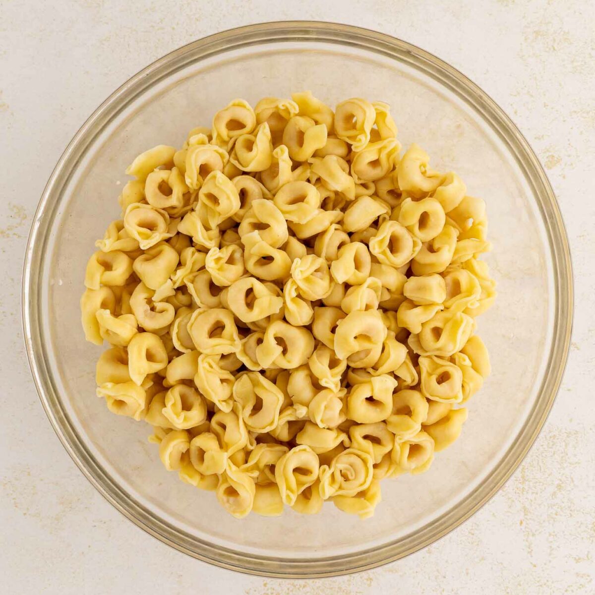 A glass bowl filled with uncooked cheese tortellini pasta sits on a light, textured surface.