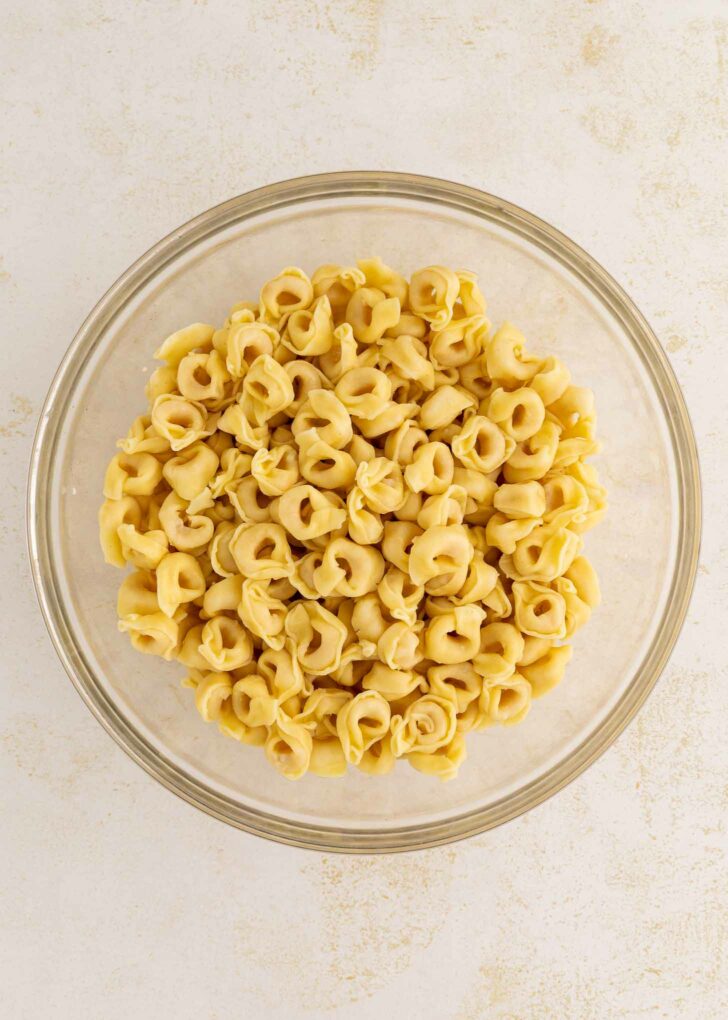 A glass bowl filled with uncooked cheese tortellini pasta sits on a light, textured surface.