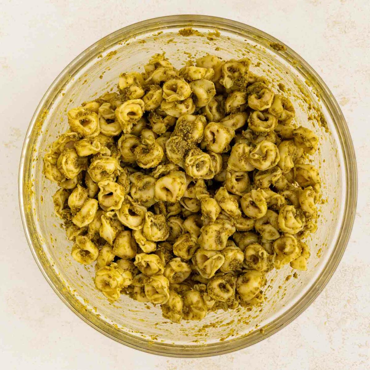 A clear glass bowl filled with tortellini pasta coated in green pesto sauce, viewed from above on a light-colored surface.