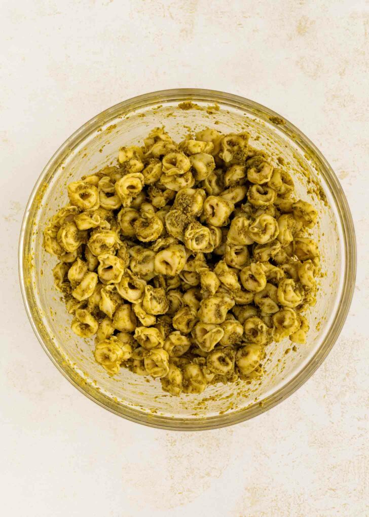 A clear glass bowl filled with tortellini pasta coated in green pesto sauce, viewed from above on a light-colored surface.