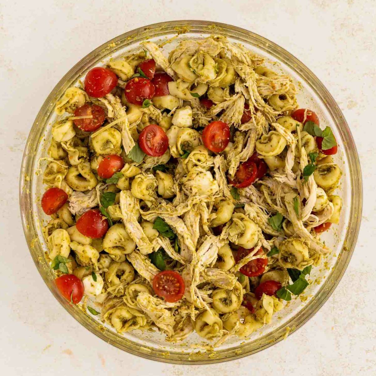 A glass bowl filled with a pasta salad made of tortellini, shredded chicken, cherry tomato halves, fresh basil, and a green pesto sauce on a light background.
