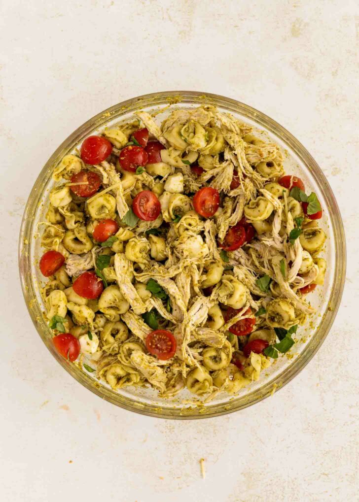 A glass bowl filled with a pasta salad made of tortellini, shredded chicken, cherry tomato halves, fresh basil, and a green pesto sauce on a light background.