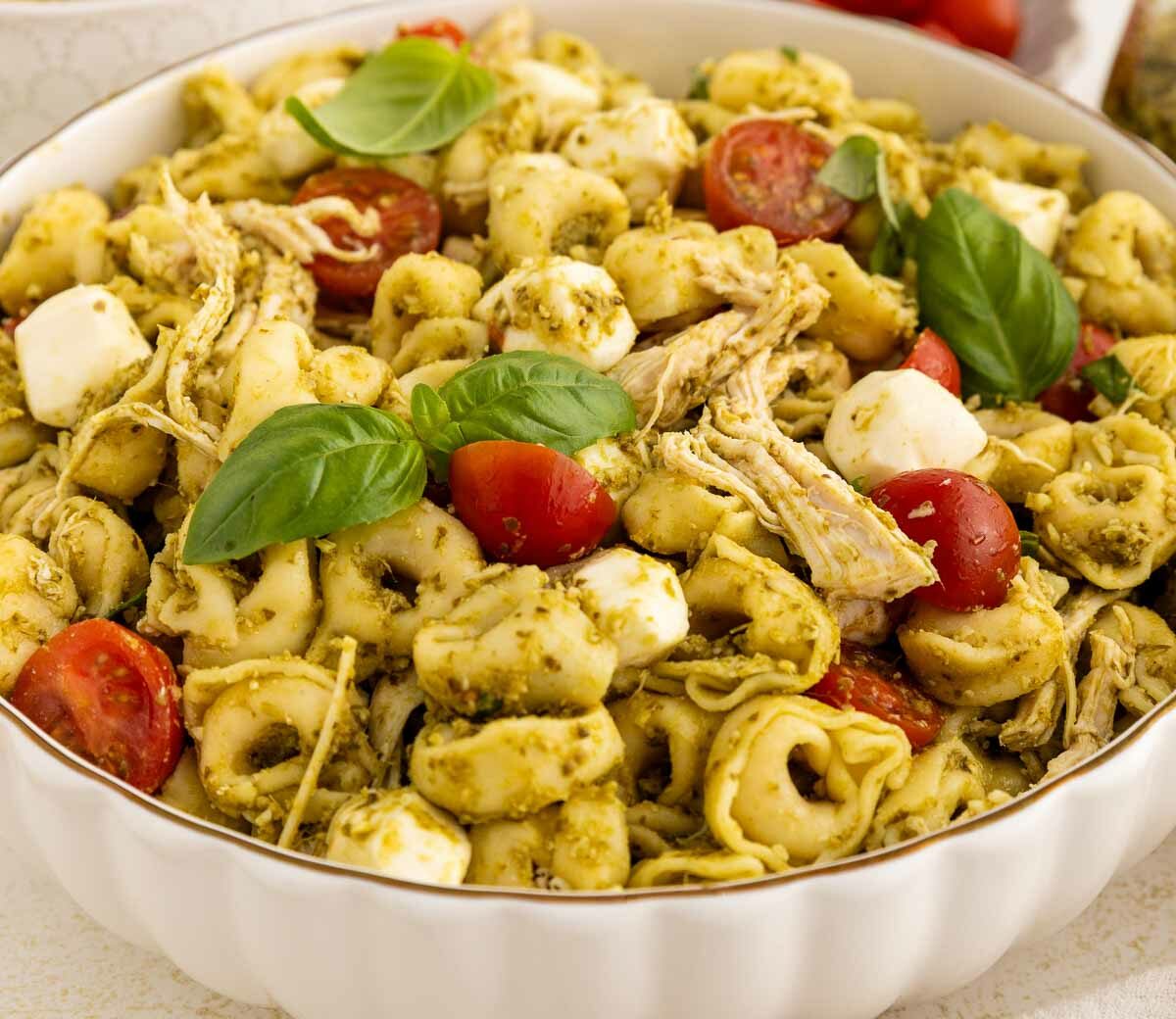 A bowl of tortellini pasta salad with shredded chicken, halved cherry tomatoes, mozzarella balls, pesto sauce, and fresh basil leaves.