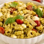 A bowl of tortellini pasta salad with shredded chicken, halved cherry tomatoes, mozzarella balls, pesto sauce, and fresh basil leaves.
