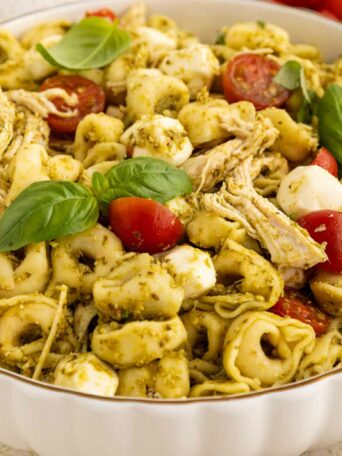 A bowl of tortellini pasta salad with shredded chicken, halved cherry tomatoes, mozzarella balls, pesto sauce, and fresh basil leaves.