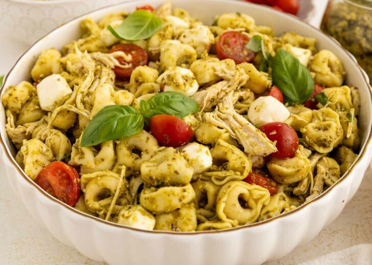 A bowl of tortellini pasta salad with shredded chicken, halved cherry tomatoes, mozzarella balls, pesto sauce, and fresh basil leaves.