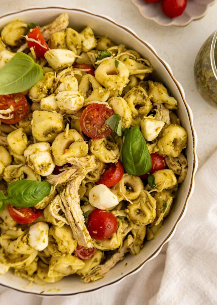 A bowl of pesto tortellini salad with shredded chicken, cherry tomatoes, mozzarella balls, and fresh basil leaves, viewed from above.
