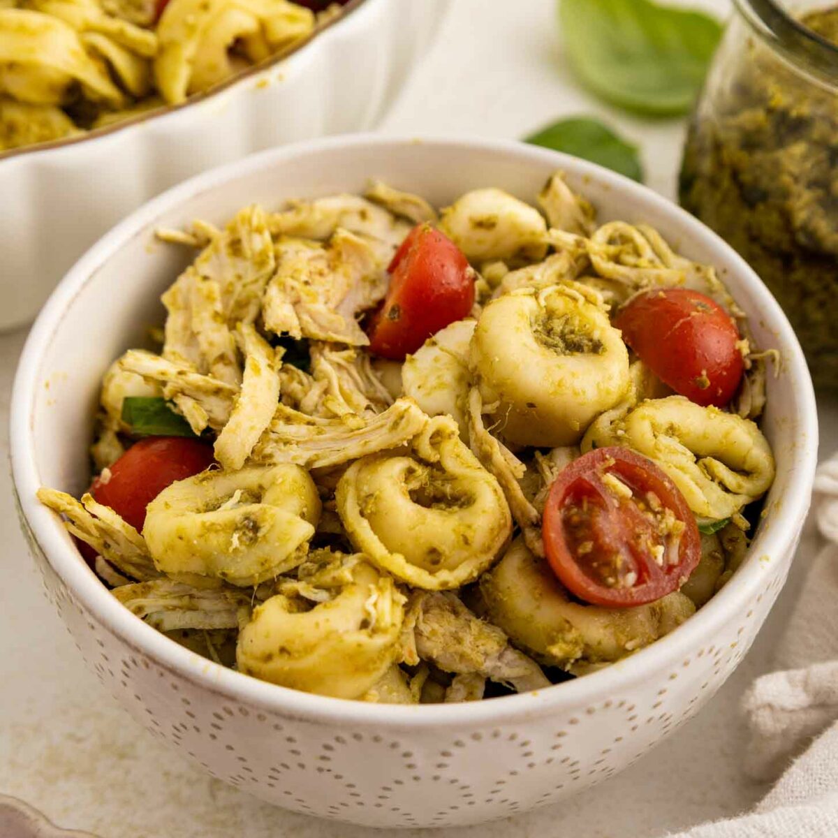 A white bowl filled with tortellini pasta, shredded chicken, halved cherry tomatoes, and pesto sauce. A jar of pesto, fresh basil leaves, and more pasta salad are visible in the background.