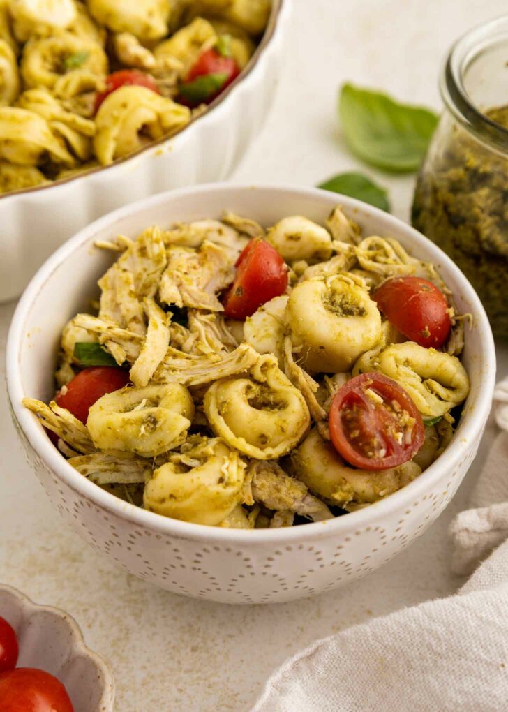 A white bowl filled with tortellini pasta, shredded chicken, halved cherry tomatoes, and pesto sauce. A jar of pesto, fresh basil leaves, and more pasta salad are visible in the background.