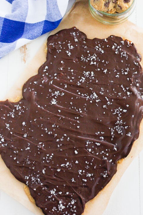 A large slab of cookie dough in a smooth layer of chocolate and sprinkled with sea salt sits on parchment paper. A blue and white checkered cloth and a small jar of cookies are visible in the background.