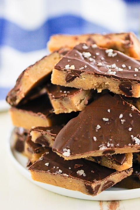 A plate stacked with pieces of chocolate-covered cookie dough, topped with flaky sea salt. The dough has a golden base and a thick chocolate layer, set against a blurred blue and white background.
