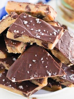 A bowl filled with pieces of cookie dough bark topped with sea salt. The dough has a thick chocolate layer and is arranged in a stacked pile on a white plate.