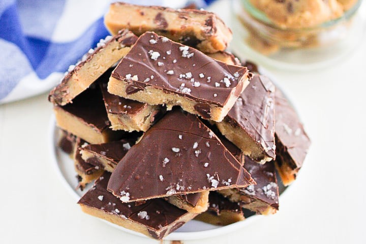 A bowl filled with pieces of cookie dough bark topped with sea salt. The dough has a thick chocolate layer and is arranged in a stacked pile on a white plate.