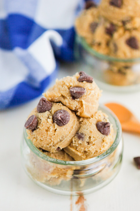 A small glass jar filled with scoops of chocolate chip cookie dough, topped with chocolate chips. A blue and white cloth and a wooden spoon are in the background.