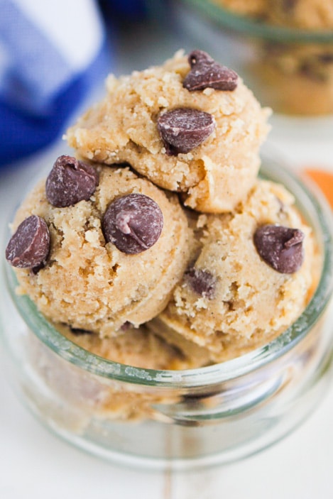 Three scoops of chocolate chip cookie dough are stacked in a small glass jar, topped with several chocolate chips. The dough looks creamy and textured, ready to be baked or eaten as is.