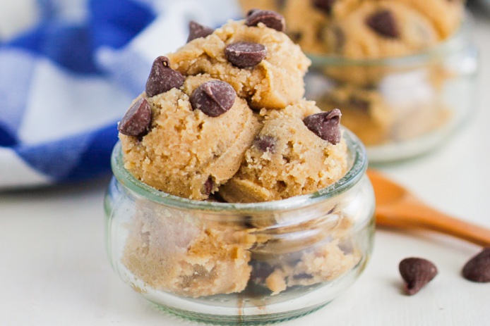 Scoops of chocolate chip cookie dough are served in a small glass jar, with visible chocolate chips on top. A wooden spoon and a blue and white cloth are in the background.