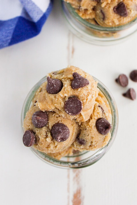 Scoops of chocolate chip cookie dough in a glass jar, viewed from above, with a few chocolate chips scattered nearby on a white surface.