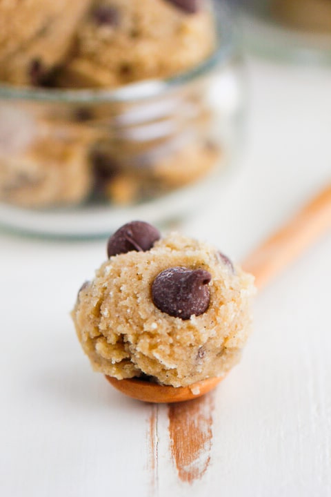 A wooden spoon holding a scoop of chocolate chip cookie dough, with visible chocolate chips, on a white surface. A bowl of more cookie dough is blurred in the background.