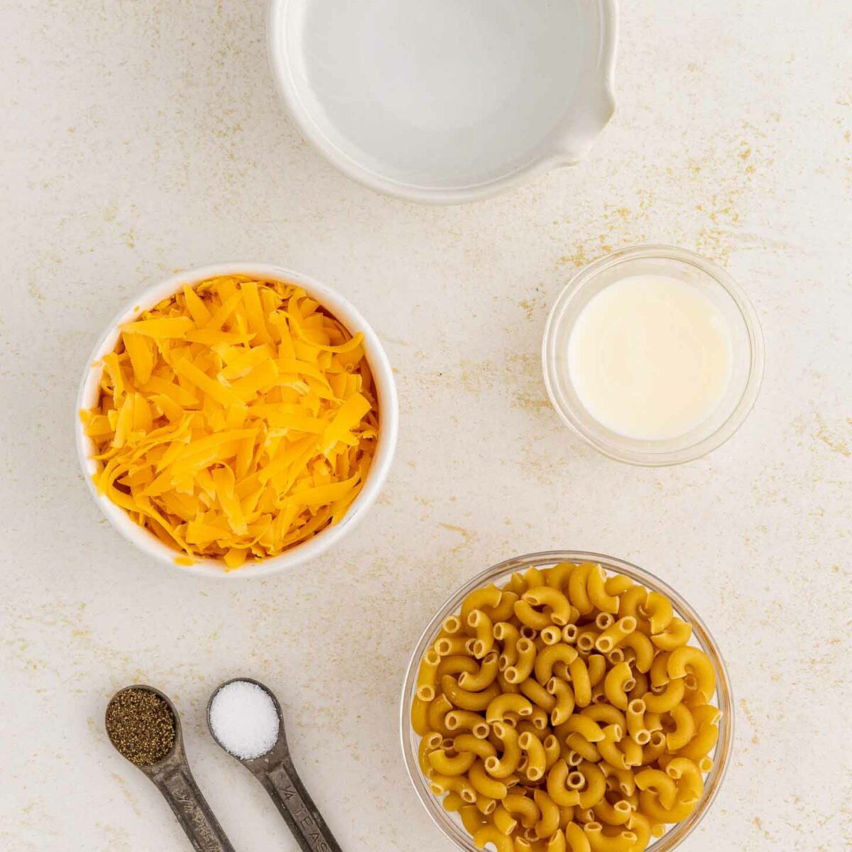 A flat lay of mac and cheese ingredients: a bowl of shredded cheddar cheese, uncooked elbow macaroni, a small bowl of cream, a bowl of water, and two measuring spoons with salt and pepper.