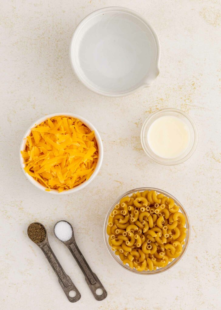 A flat lay of mac and cheese ingredients: a bowl of shredded cheddar cheese, uncooked elbow macaroni, a small bowl of cream, a bowl of water, and two measuring spoons with salt and pepper.