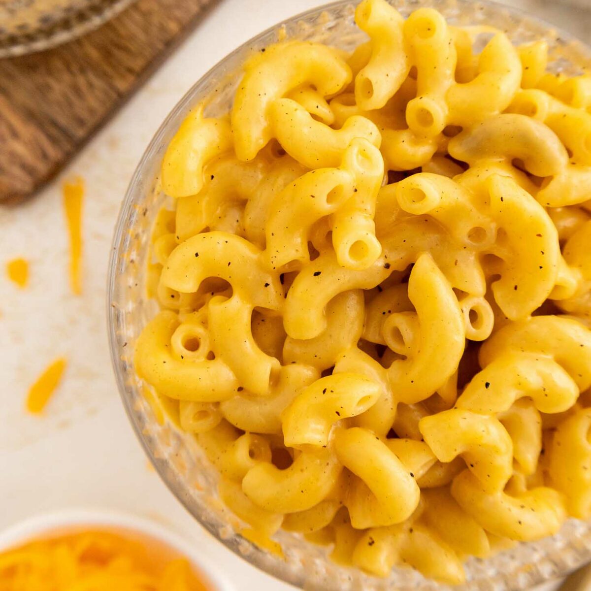 A close-up of a bowl filled with creamy macaroni and cheese, with elbow-shaped pasta coated in a rich, yellow cheese sauce. Shredded cheese and a cloth napkin are visible in the background.