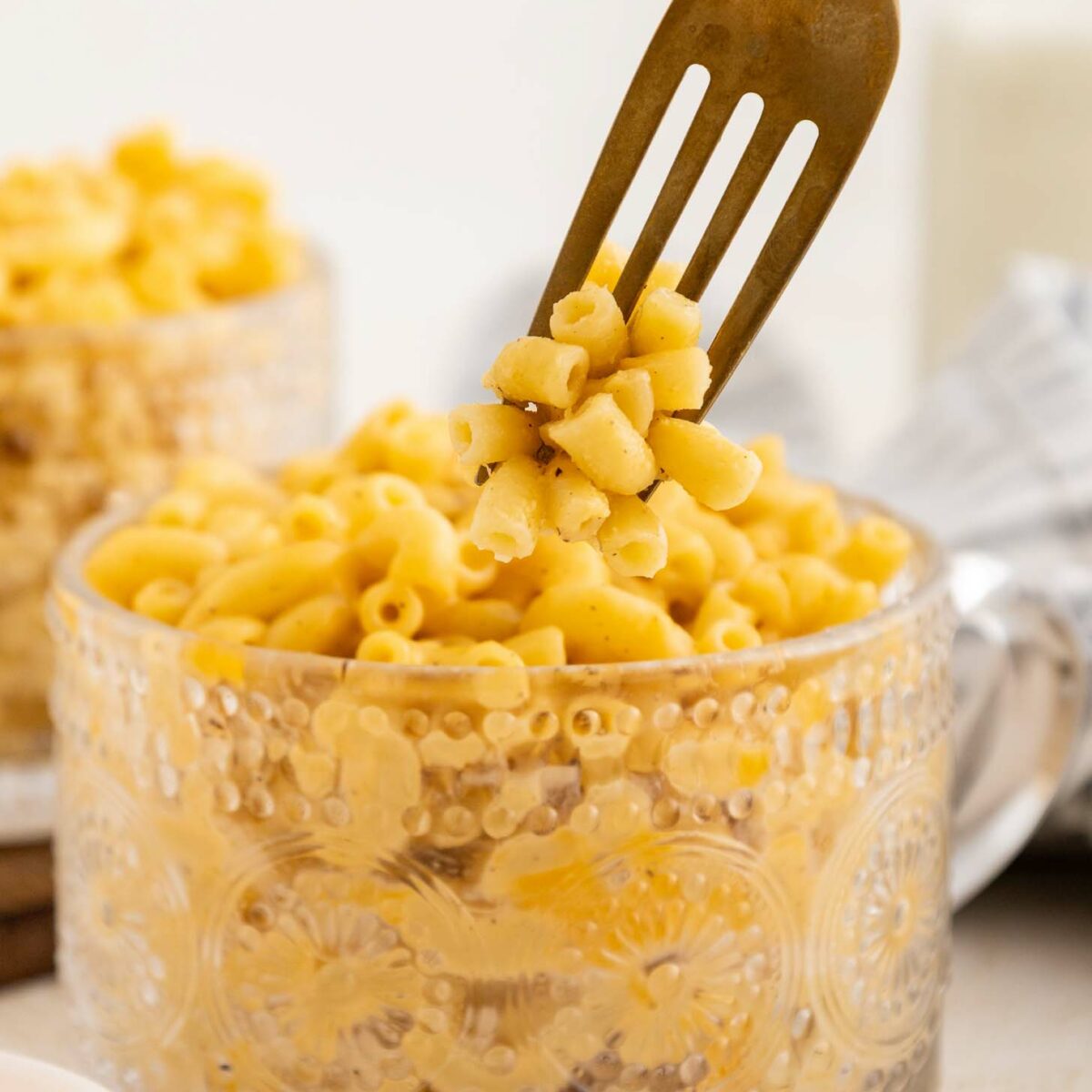 A fork lifts a portion of creamy macaroni and cheese from a decorative glass mug filled with pasta. Another cup of mac and cheese and a milk bottle are blurred in the background.