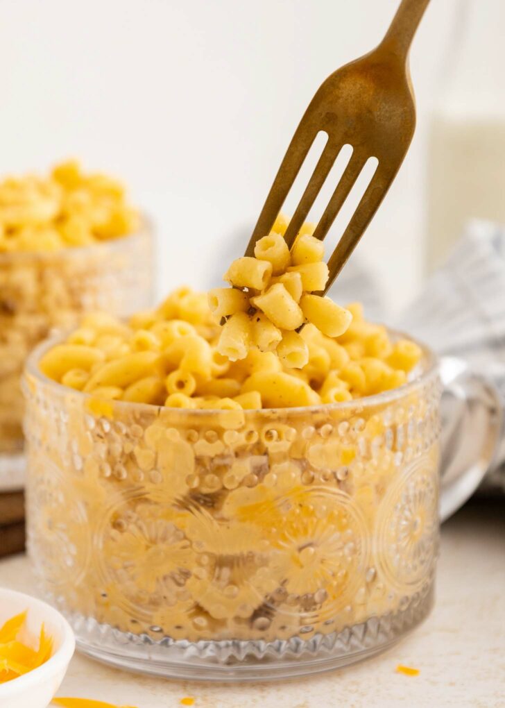 A fork lifts a portion of creamy macaroni and cheese from a decorative glass mug filled with pasta. Another cup of mac and cheese and a milk bottle are blurred in the background.