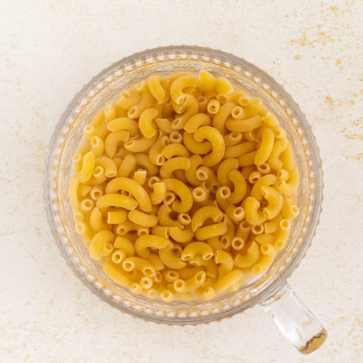 A clear glass measuring cup filled with uncooked elbow macaroni noodles, viewed from above, on a light textured surface.