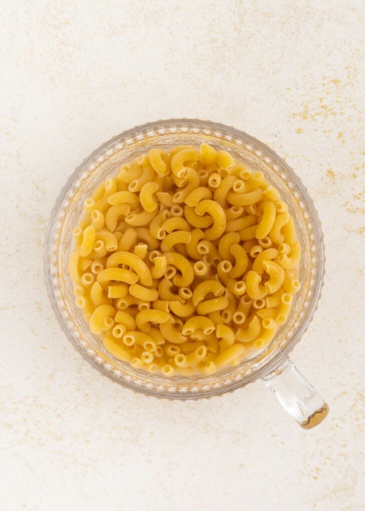 A clear glass measuring cup filled with uncooked elbow macaroni noodles, viewed from above, on a light textured surface.