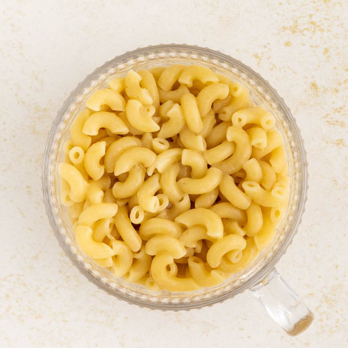 A clear glass mug filled with cooked elbow macaroni and creamy cheese sauce sits on a light, textured surface.