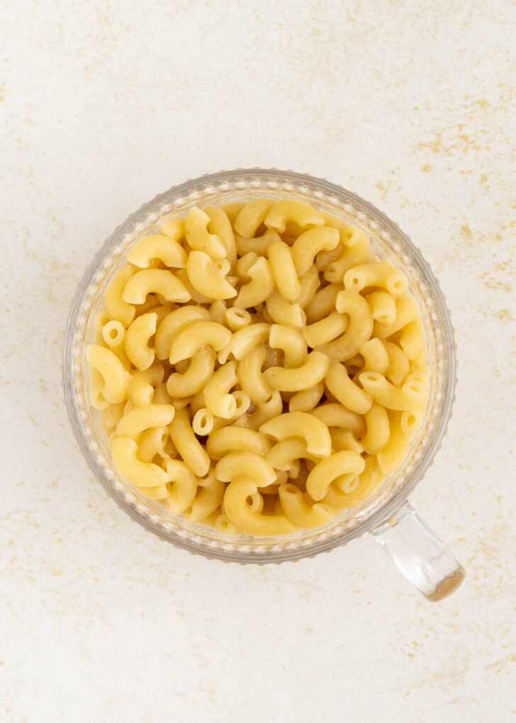 A clear glass mug filled with cooked elbow macaroni and creamy cheese sauce sits on a light, textured surface.