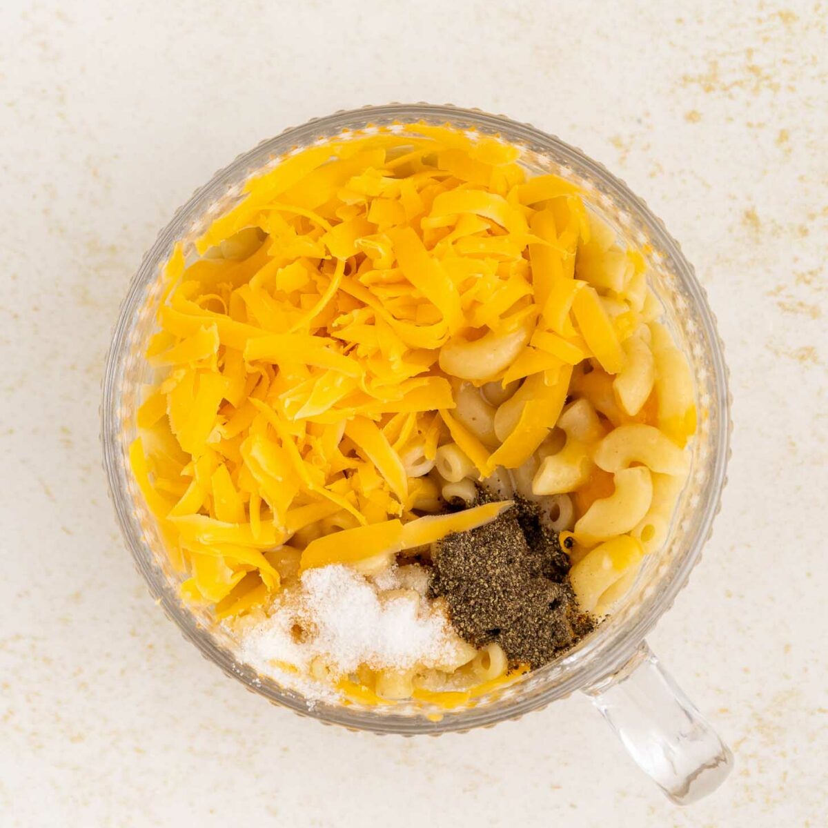 A clear glass mug filled with cooked macaroni, shredded cheddar cheese, ground black pepper, and salt on a light, speckled surface.
