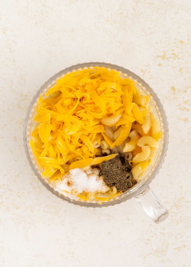 A clear glass mug filled with cooked macaroni, shredded cheddar cheese, ground black pepper, and salt on a light, speckled surface.
