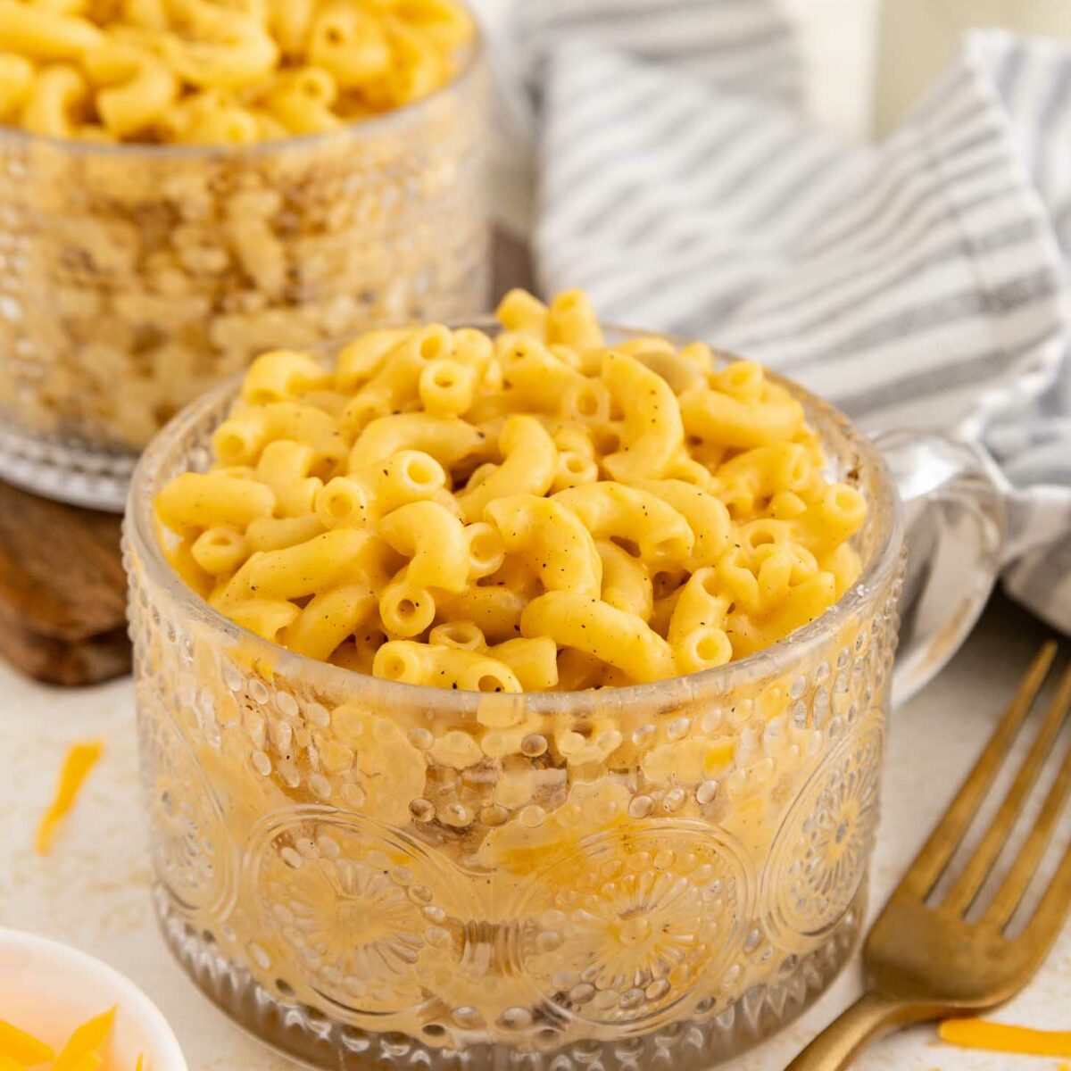 A clear glass mug filled with creamy macaroni and cheese sits on a table next to a gold fork, a striped gray napkin, and another mug of macaroni in the background. Shredded cheese is in a small bowl nearby.