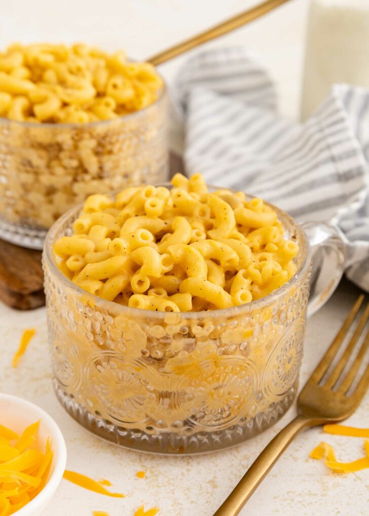 A clear glass mug filled with creamy macaroni and cheese sits on a table next to a gold fork, a striped gray napkin, and another mug of macaroni in the background. Shredded cheese is in a small bowl nearby.