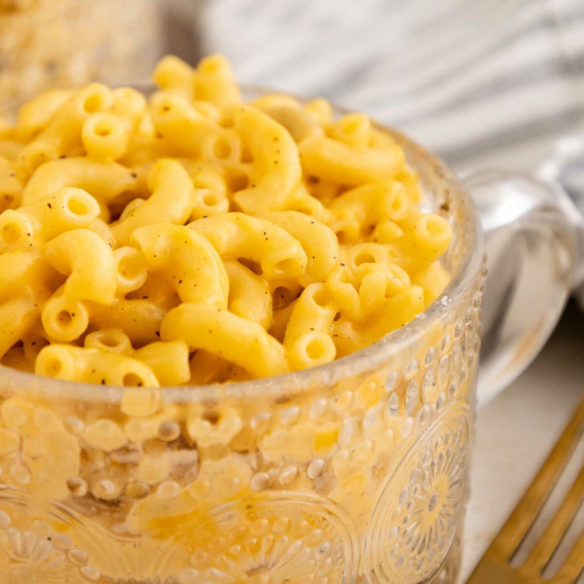 A clear, patterned glass cup filled with creamy macaroni and cheese sits on a table beside a gold fork and a striped cloth napkin.