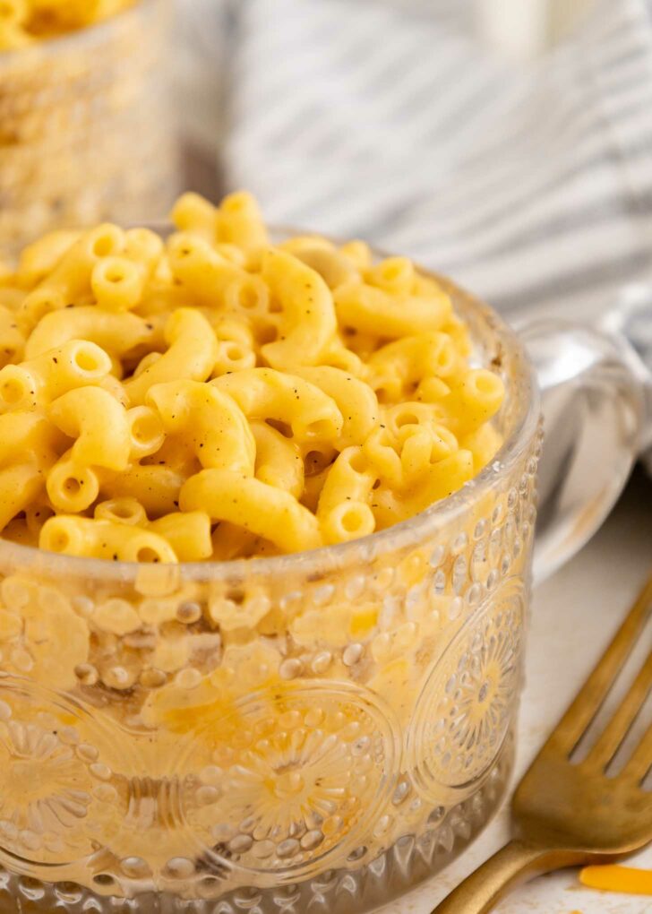 A clear, patterned glass cup filled with creamy macaroni and cheese sits on a table beside a gold fork and a striped cloth napkin.