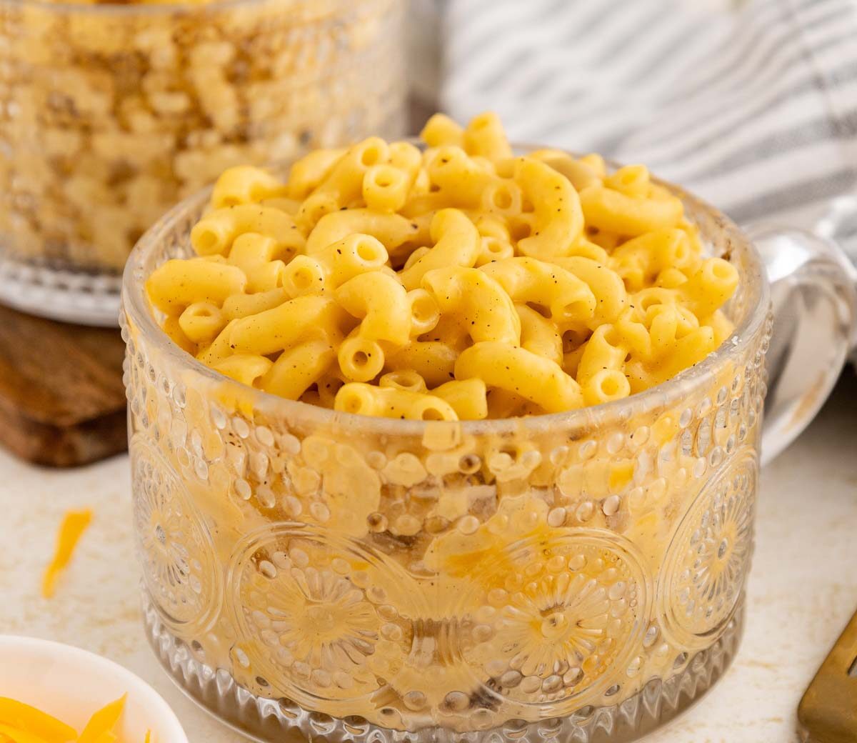 A clear glass mug filled with creamy macaroni and cheese sits on a table next to a striped cloth napkin and a fork, with a bowl of shredded cheese partially visible nearby.