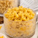 A clear glass mug filled with creamy macaroni and cheese sits on a table next to a striped cloth napkin and a fork, with a bowl of shredded cheese partially visible nearby.
