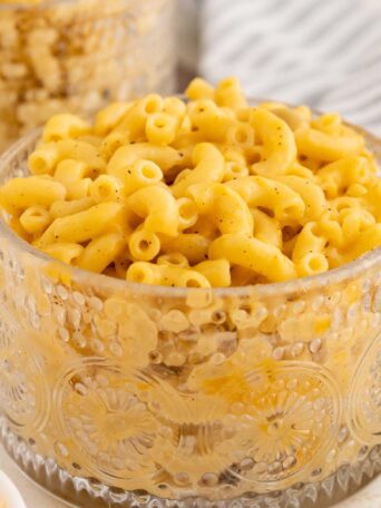 A clear glass mug filled with creamy macaroni and cheese sits on a table next to a striped cloth napkin and a fork, with a bowl of shredded cheese partially visible nearby.