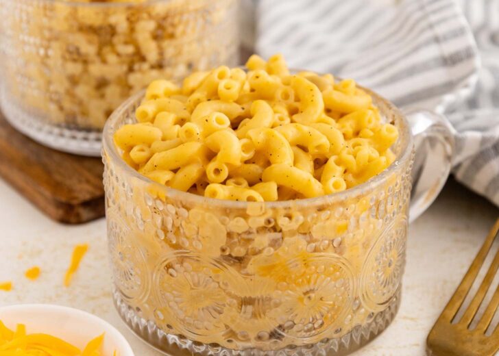 A clear glass mug filled with creamy macaroni and cheese sits on a table next to a striped cloth napkin and a fork, with a bowl of shredded cheese partially visible nearby.