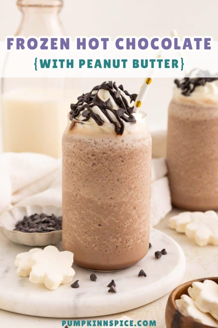 A mason jar filled with frozen hot chocolate topped with whipped cream, chocolate drizzle, and chocolate chips, with a paper straw. Peanut butter and milk are in the background, with marshmallow flowers and chocolate chips nearby.
